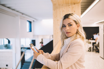 Fototapeta na wymiar Portrait of confident female lawyer standing in modern workshop with smartphone device in hand and looking at camera, caucasian businesswoman using cellular phone during working time indoors