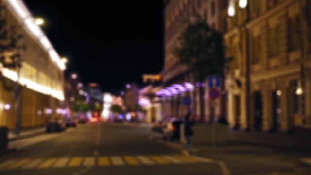 Defocused view of the city street. Night time. People walking on the sidewalk. Pedestrians crossing a road. Illuminated facades of houses and parked cars in the background. Ultra HD