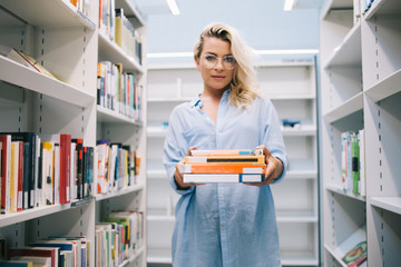 Serious young woman in casual wear standing near bookshelves holding literature for education in college, caucasian female librarian in optical spectacles offering books for reading on leisure