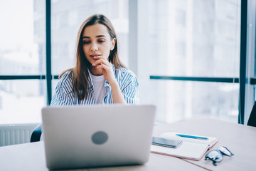 Pensive female woman watching video online on netbook during working process for publicating...