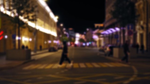 Defocused view of the city street. Night. People crossing pedestrian crossing. Pedestrians walking on the sidewalk. Cars driving on the road. Illuminated facades of houses in the background. Ultra HD