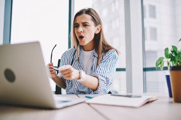 Emotional woman stressed while watching video on laptop computer sitting in office interior, shocked female pointing on netbook irritated about application failure during working process at desktop.