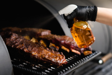 Professional kitchen appliance. Closeup of beef and pork ribs cooked in BBQ smoker. Cropped shot of chef spraying grilled meat.