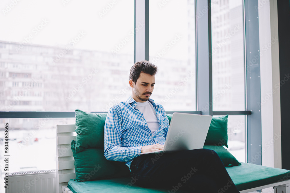 Poster concentrated young man dressed in casual wear downloading media file from internet website while cre