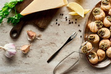 top view of delicious cooked escargots with lemon slices, Parmesan, garlic, parsley, black peppercorn and tweezers on stone background