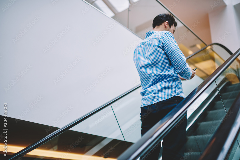 Poster back view of millennial hipster guy standing on escalator in shopping center and using public wirele