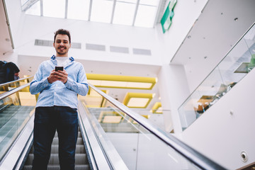 Half length portrait of cheerful man looking at camera while going down on modern escalator on publicity area holding smartphone for checking balance on credit card via application before shopping