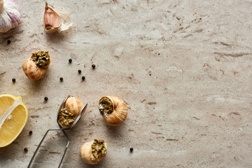 top view of delicious cooked escargots with lemon, garlic, black peppercorn and tweezers on stone background