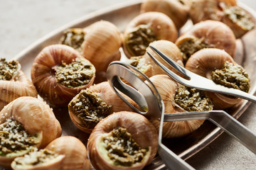 close up view of delicious escargots with tweezers on plate on stone background