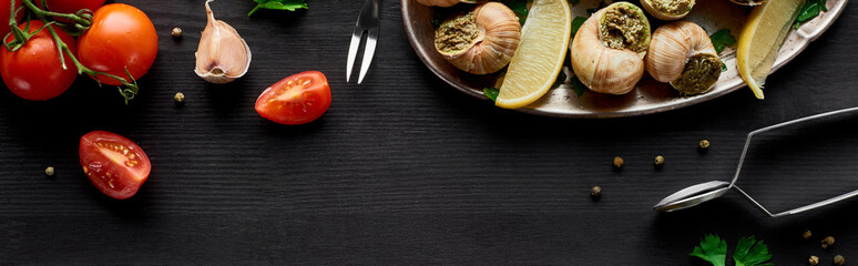 top view of delicious cooked escargots with lemon slices on black wooden table with ingredients, panoramic shot