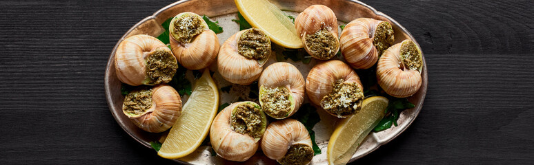 top view of delicious cooked escargots with lemon on black wooden table, panoramic shot