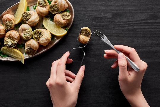 cropped view of woman eating delicious escargots with lemon on black wooden table