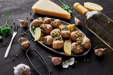 Delicious cooked escargots with lemon slices on black wooden table with spices, Parmesan and white wine