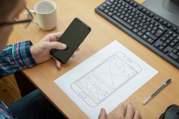 Designer sketches the layout of a responsive website or app and tests it on a mobile phone. Close up, work desk with computer beside.