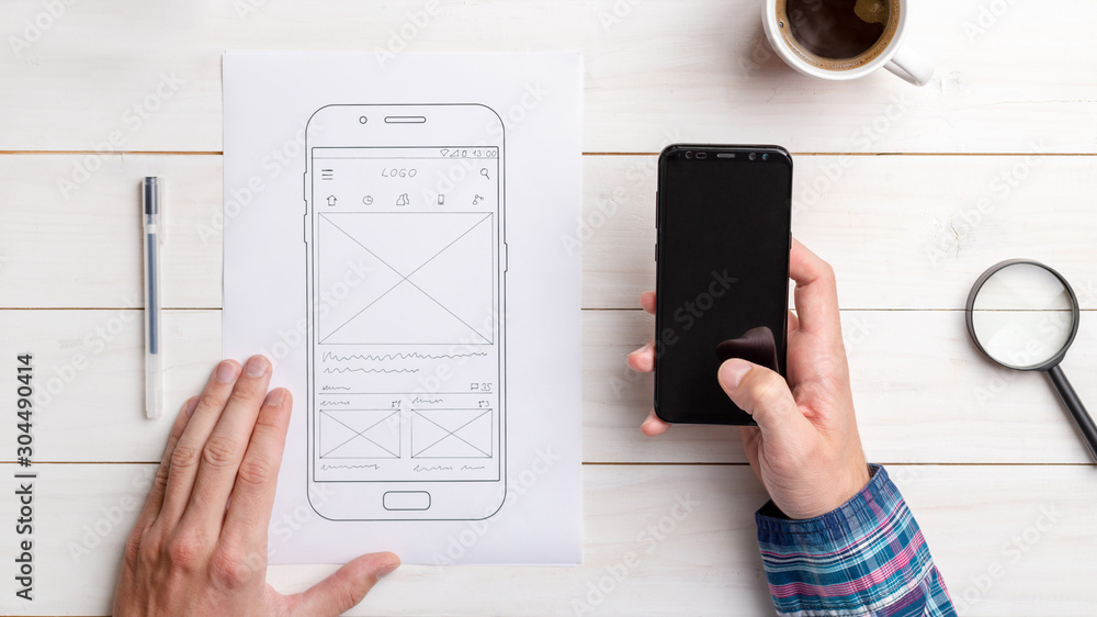Fototapete Designer tests the user interface and user experience on a mobile phone. Wireframe beside with mobile phone with a sketched app. The concept of designing software, apps and websites.