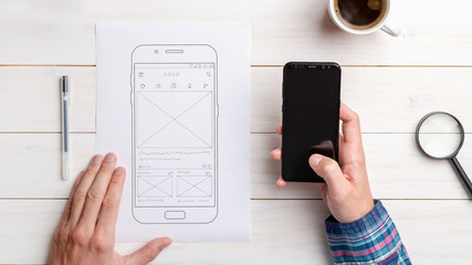 Designer tests the user interface and user experience on a mobile phone. Wireframe beside with mobile phone with a sketched app. The concept of designing software, apps and websites.