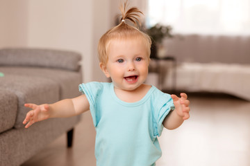 healthy baby girl in a blue bodysuit in a room next to a gray sofa is learning to walk. the comfort of the home. the child smiles