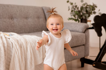 healthy baby girl in a white bodysuit in a room next to a gray sofa is learning to walk. the comfort of the home. the child smiles