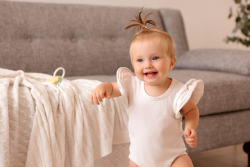 healthy baby girl in a white bodysuit in a room next to a gray sofa is learning to walk. the comfort of the home. the child smiles