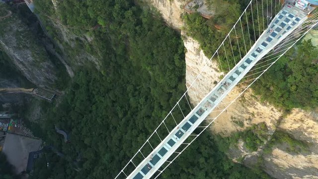 Tilting aerial view of tourists crossing spectacular glass walkway (Zhangjiajie Glass Bridge), steep mountain valley in famous national park, natural landscapes and popular travel destinations in Chin