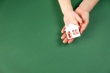 Top view of female and child hands holding white paper house on green background. Family home and...