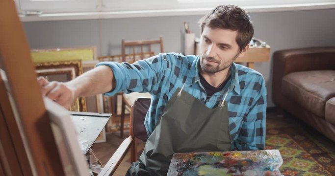 Attractive bearded male artist painting picture in cozy studio and looking to camera, portrait