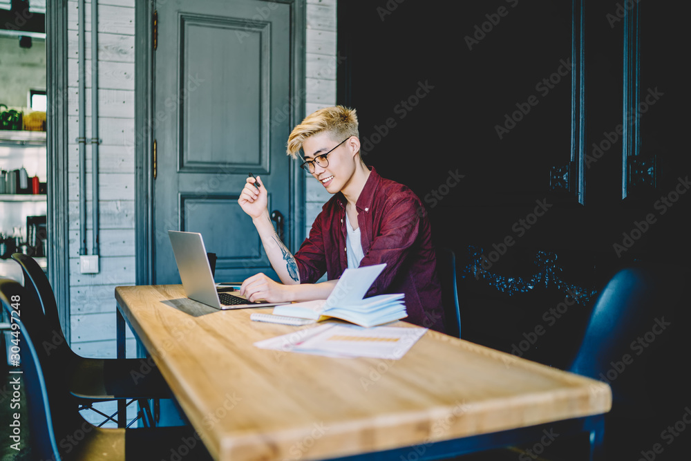 Poster positive japanese young man sitting with laptop and working remotely in coworking space, hipster guy