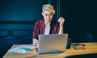 Wondered chinese young man working on freelance and has problem with software on modern laptop device.Confused hipster guy watching webinar online on computer device using wireless internet connection