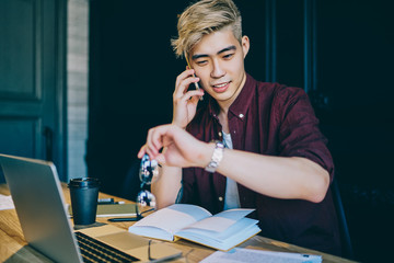 Fototapeta na wymiar Blonde positive chinese young man checking time on smartwatch while calling on smartphone during working process at laptop.Stylish japanese student looking at wristwatch and talking on cellular