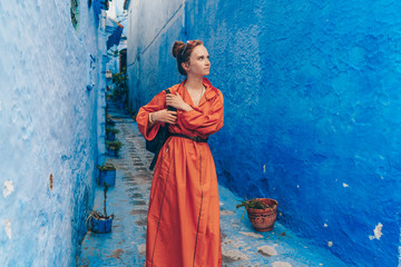 a tourist in a long bright orange dress with a backpack walks through the blue city of Morocco