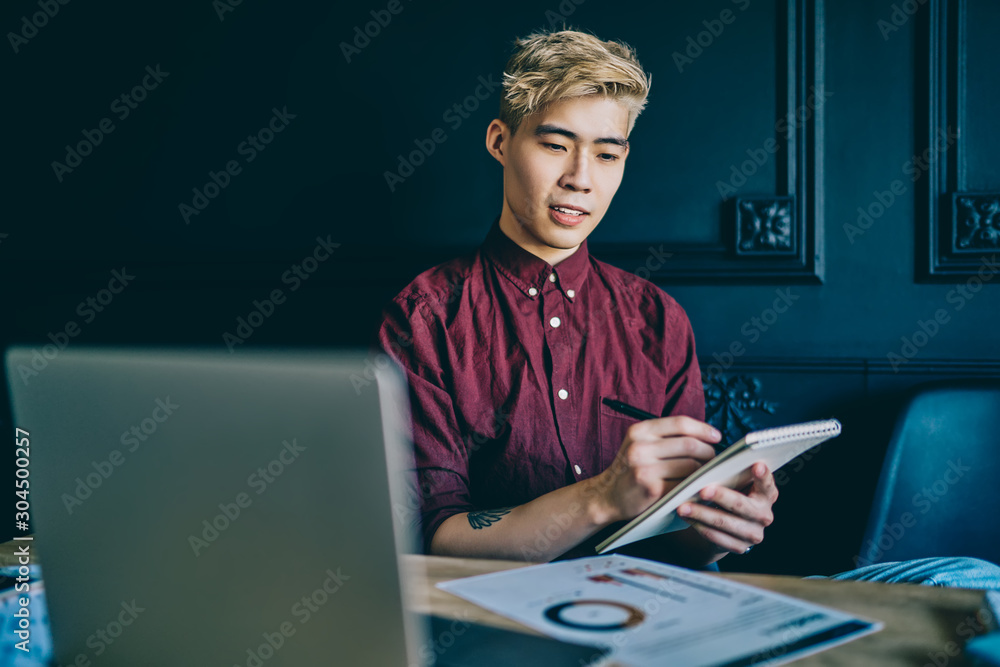 Poster pensive blonde japanese hipster guy holding notepad in hand and writing down checklist sitting at la