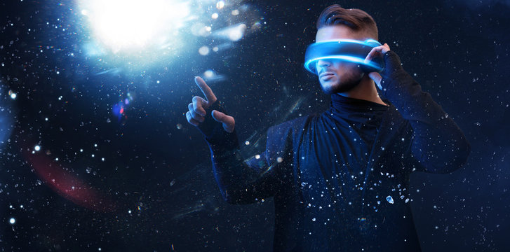 Beautiful young man using VR helmet while touching air in colorful neon lights. Guy in glasses of virtual reality with blue backlight over dark magic universe background.  - Powered by Adobe
