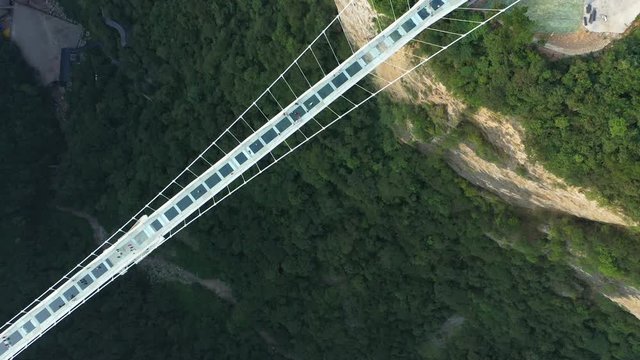 Overhead drone shot of tourists walking over glass suspension bridge, looking around 300 meters down on beautiful mountain valley, landmark travel destination in China