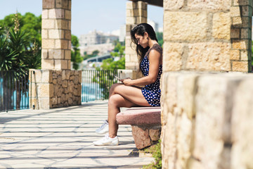 Beautiful hispanic woman in blue dress sitting on a bench while using a smartphone