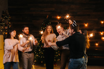 Cheerful friends are dancing with champagne glasses and with Bengali lights, celebrating New Year. Christmas tree with garland in background. Friends are celebrating Christmas Eve.