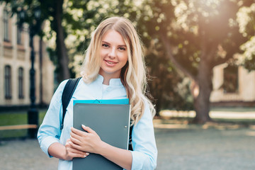 Student girl is smiling and holding a folder and a notebook in her hands at university background....
