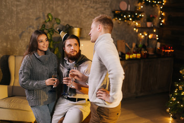 Young man talks to couple in celebrating new years eve party in festively decorated room holding glasses of champagne in hand. Christmas tree in background.