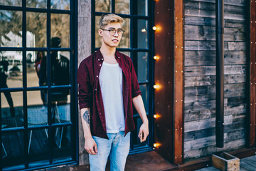 Young handsome hipster guy in stylish optical spectacles for provide eyes protection dressed in trendy apparel standing outdoors on cozy urban setting and looking forward during leisure time