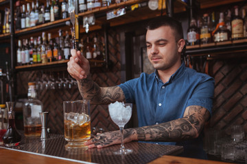 Handsome tattooed bartender smiling, mixing alcohol, preparing cocktails