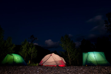 Three tents glowing from the inside on a night forest background. Dark blue sky, trees silhouettes, stone soil. Copy space. The concept of travel, leisure and wild tourism.