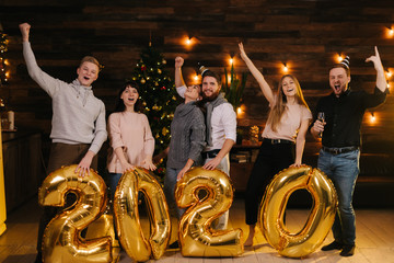 Fototapeta na wymiar Bunch of happy friends are holding inflatable foil numerals 2020. Group of cheerful young man and woman are celebrating New Year. Christmas tree with garland and festive light in background.