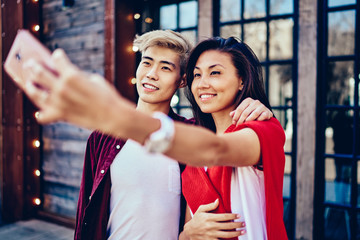 Happy chinese couple in love standing together outdoors on background with modern cafe and making photo on front camera of smartphone.Positive asian friends taking selfie picture on cellular