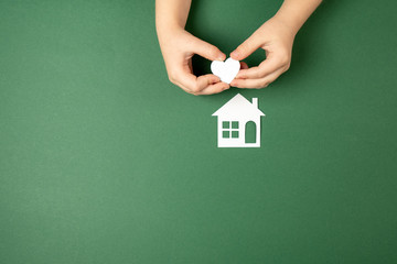 Family home and real estate concept. Close up of child hands holding white paper house and heart on green background. Flat lay, copy space