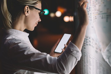 Digital monitor with public transport subway map. Female standing at big display with smartphone in hand. Young woman touching with finger screen while using train schedule application on mobile phone