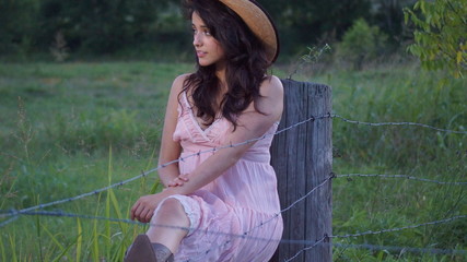 Beautiful woman sitting on a fence staring down at the ground with her hands on her hat