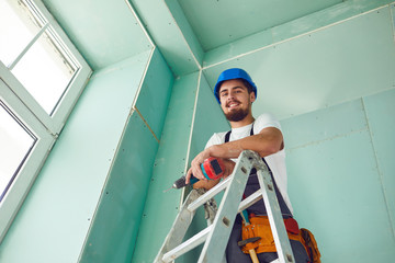 A builder standing on a ladder installs drywall at a construction site
