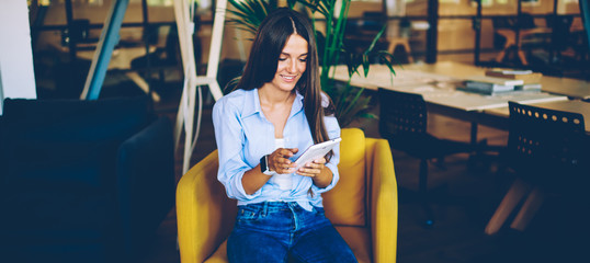 Cheerful hipster girl watching comedy show on website using digital tablet and free wireless connection, smiling woman browsing internet on modern touch pad while sitting in comfortable chair indoors