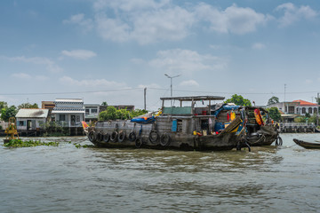 Fototapeta na wymiar Cai Be, Mekong Delta, Vietnam - March 13, 2019: Along Kinh 28 canal. Two old gray wooden barges with black car tires on brown water under blue cloudscape. Houses on shore. Household items add colors.