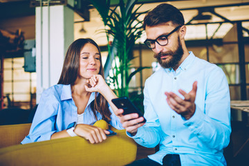 Positive smiling hipster girl listening male friend sitting near and searching information in internet via smartphone device, happy woman enjoying time with guy indoors, concept of technology