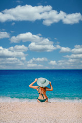 Fototapeta na wymiar Young red-haired woman in a hat and a bathing suit sits on the beach on a sunny day, rear view